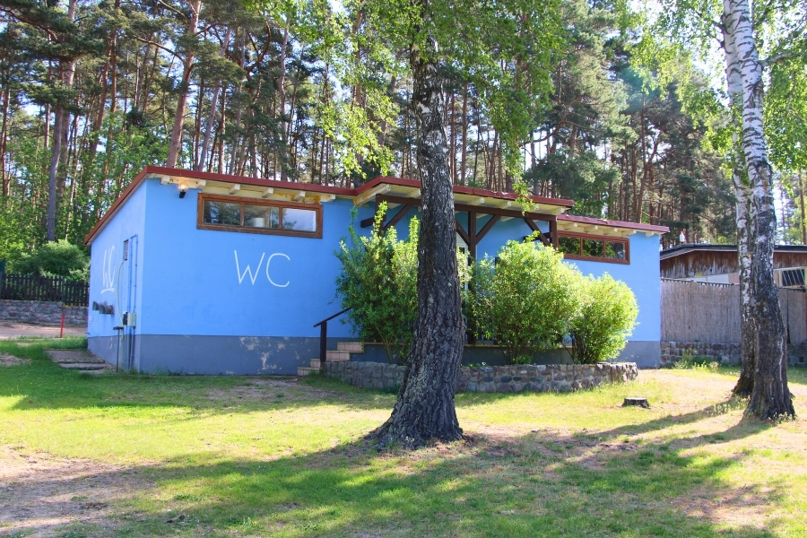 Ein leuchtend blau gestrichenes, einstöckiges Gebäude mit der großen weißen Aufschrift „WC“ an der Fassade steht auf einer Rasenfläche. Ein kurzer Treppenaufgang führt zu einem Eingangsbereich mit hölzernem Vordach. Das Gebäude ist im Hintergrund von einem dichten Kiefernwald umgeben, während im rechten Vordergrund der dicke, weiß-schwarze Stamm einer Birke steht.