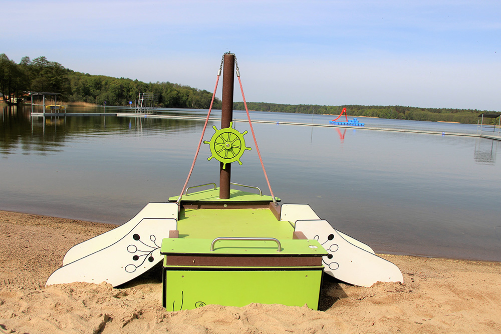 Ein buntes Spielgerät im Design eines grünen Bootes steht direkt im Sandstrand des Strandbads Wolletzsee. Das Boot verfügt über einen Mast mit einem grünen Steuerrad und Seilen, während im Hintergrund das weite Wasser des Sees, Steganlagen und das bewaldete Ufer zu sehen sind.