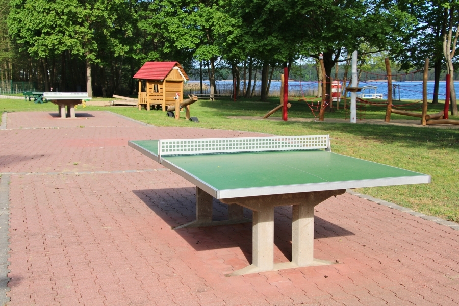 Eine robuste Outdoor-Tischtennisplatte mit grüner Spielfläche und festem, durchbrochenem Netz steht im Vordergrund auf einem rot gepflasterten Platz. Im grünen Hintergrund befinden sich unter großen Laubbäumen eine zweite Tischtennisplatte, ein kleines Holzspielhaus mit rotem Dach sowie ein Klettergerüst aus Holz. Hinter dem Spiel- und Freizeitbereich ist das blaue Wasser eines Sees zu erkennen.
