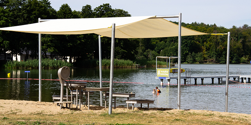 Ein schattiger Wasserspielplatz mit Tischen und spielgeräten steht direkt am Sandstrand des Strandbads Wolletzsee unter einem großen, hellen Sonnensegel. Im Hintergrund sind das Wasser des Sees, ein Steg und das bewaldete Ufer zu sehen.