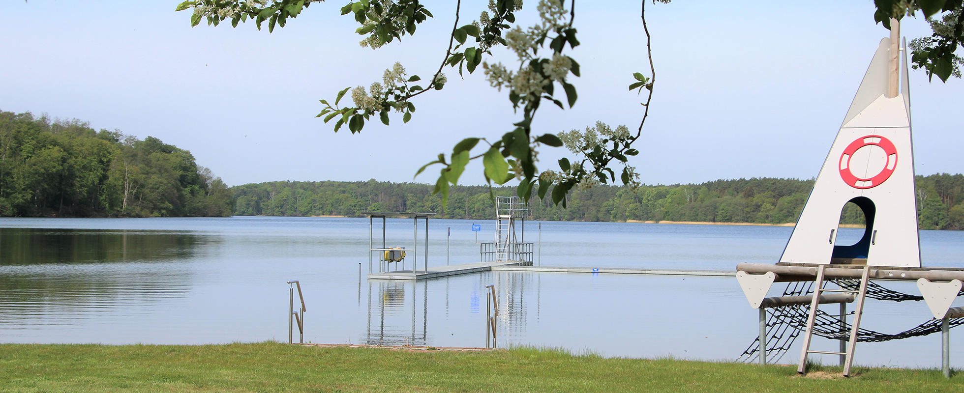 Ein Blick über eine grüne Wiese auf den weiten Wolletzsee im Strandbad Wolletzsee. Im rechten Vordergrund steht ein hölzernes Klettergerüst im Design eines Segelbootes mit einer Strickleiter und einem aufgemalten Rettungsring. Im Mittelgrund ragt eine lange Steganlage mit Sprungturm in den See, während im Hintergrund das dichte, bewaldete Ufer zu sehen ist.