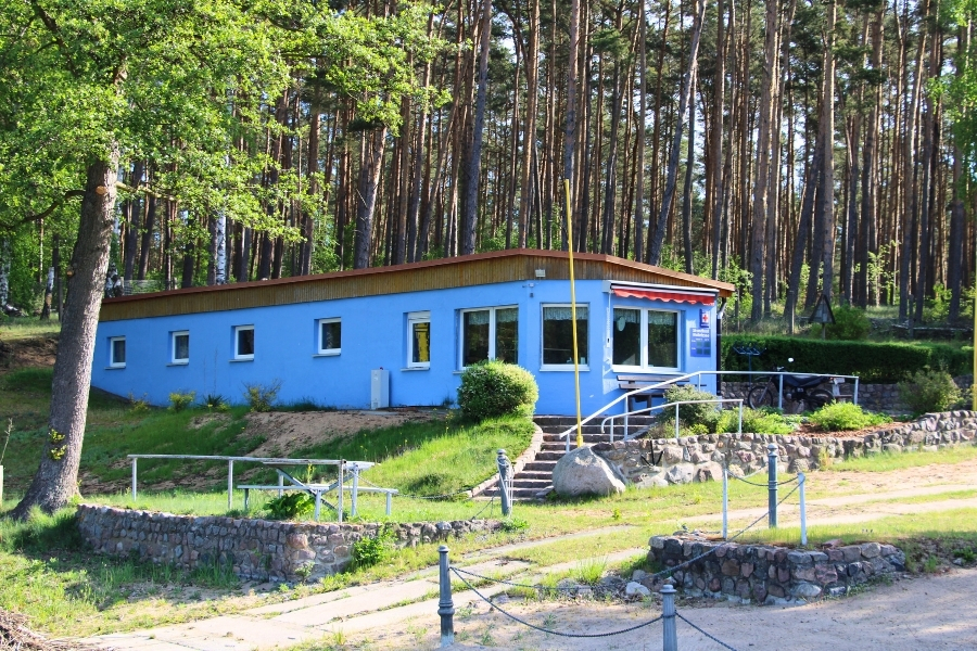 Ein langgestrecktes, hellblaues Gebäude, das leicht erhöht vor einem dichten Kiefernwald steht und durch Natursteinmauern und Treppen eingefasst ist.