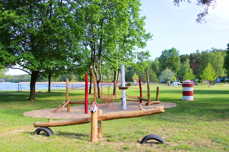 Ein Holzspielplatz mit Klettergerüst und Wippe auf einer Sandfläche, gelegen auf einer großen Wiese mit Bäumen nahe einem Seeufer.