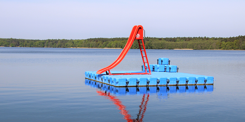 Eine rote Wasserrutsche auf einer schwimmenden, blauen Pontonplattform in einem ruhigen See, umgeben von einem bewaldeten Ufer.